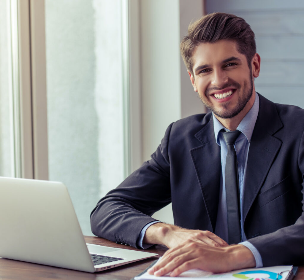 Portrait of handsome young businessman in formal suit using a laptop, looking at camera and smiling while working in office